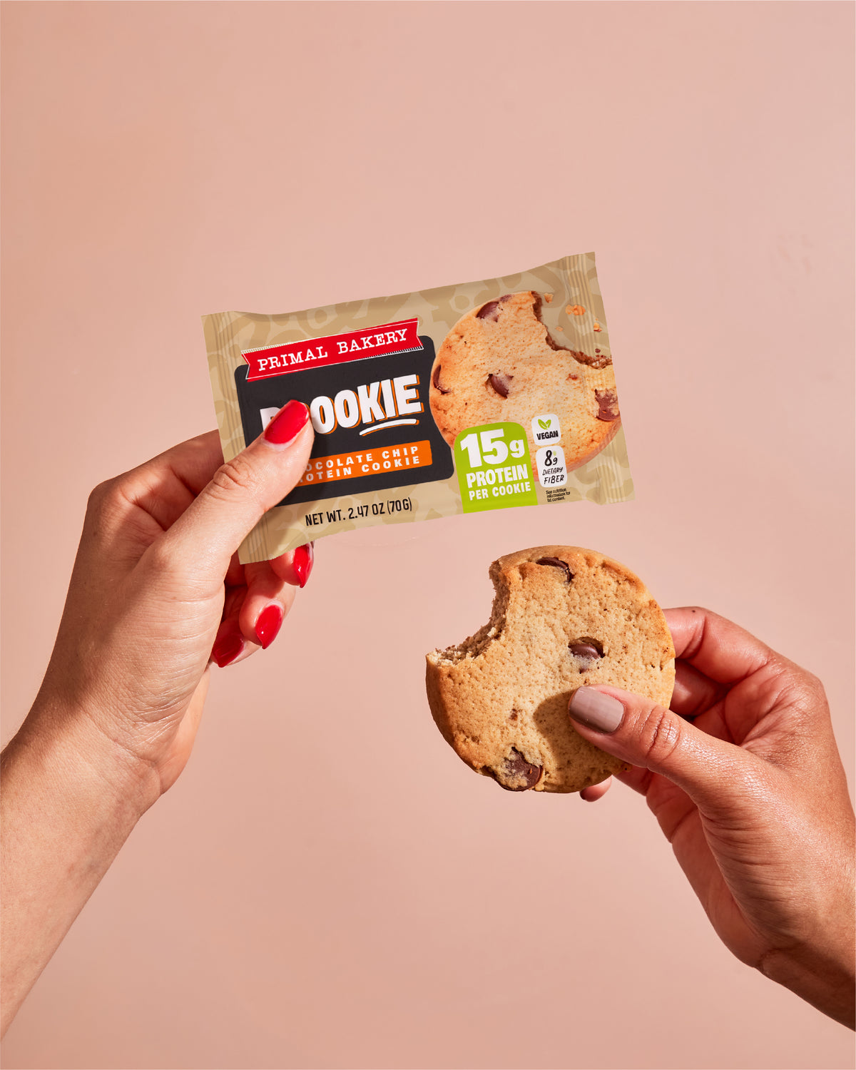 Hands holding a Primal Bakery Chocolate Chip Prookie package and a partially eaten protein cookie, with 15g protein and 8g dietary fiber per cookie