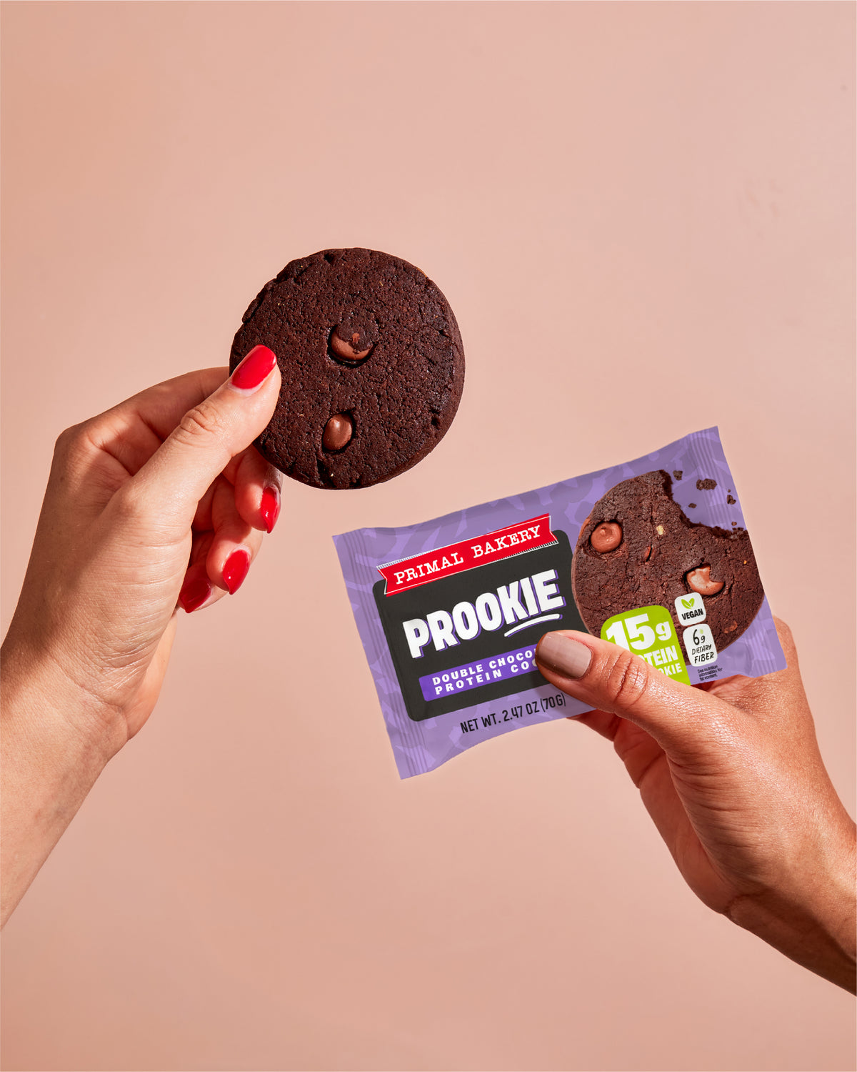 Hands holding a Primal Bakery Double Chocolate Prookie package and chocolate protein cookie, with 15g protein and 6g dietary fiber per cookie