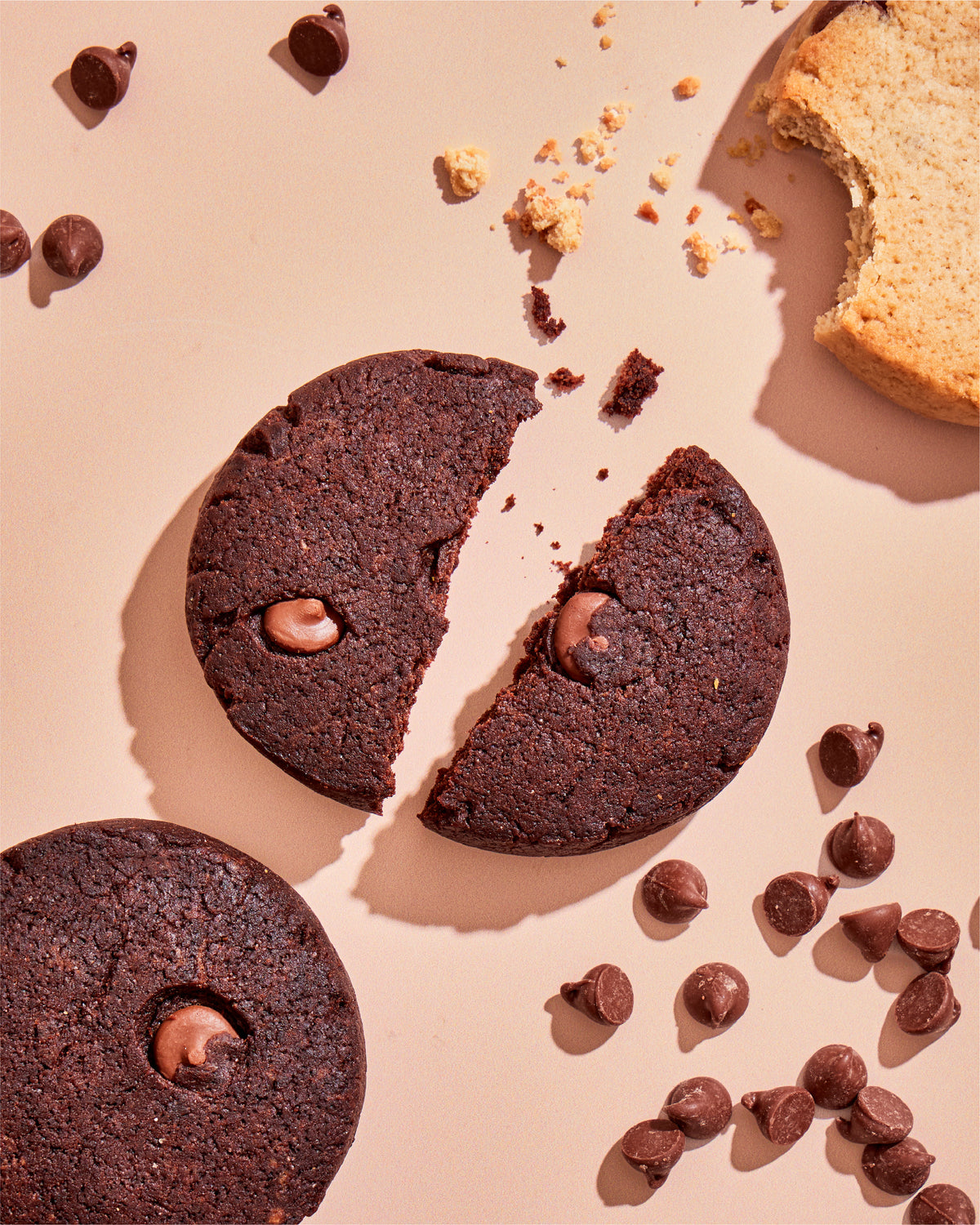 Primal Bakery Double Chocolate Prookies with one cookie broken in half and chocolate chips scattered on a pink background