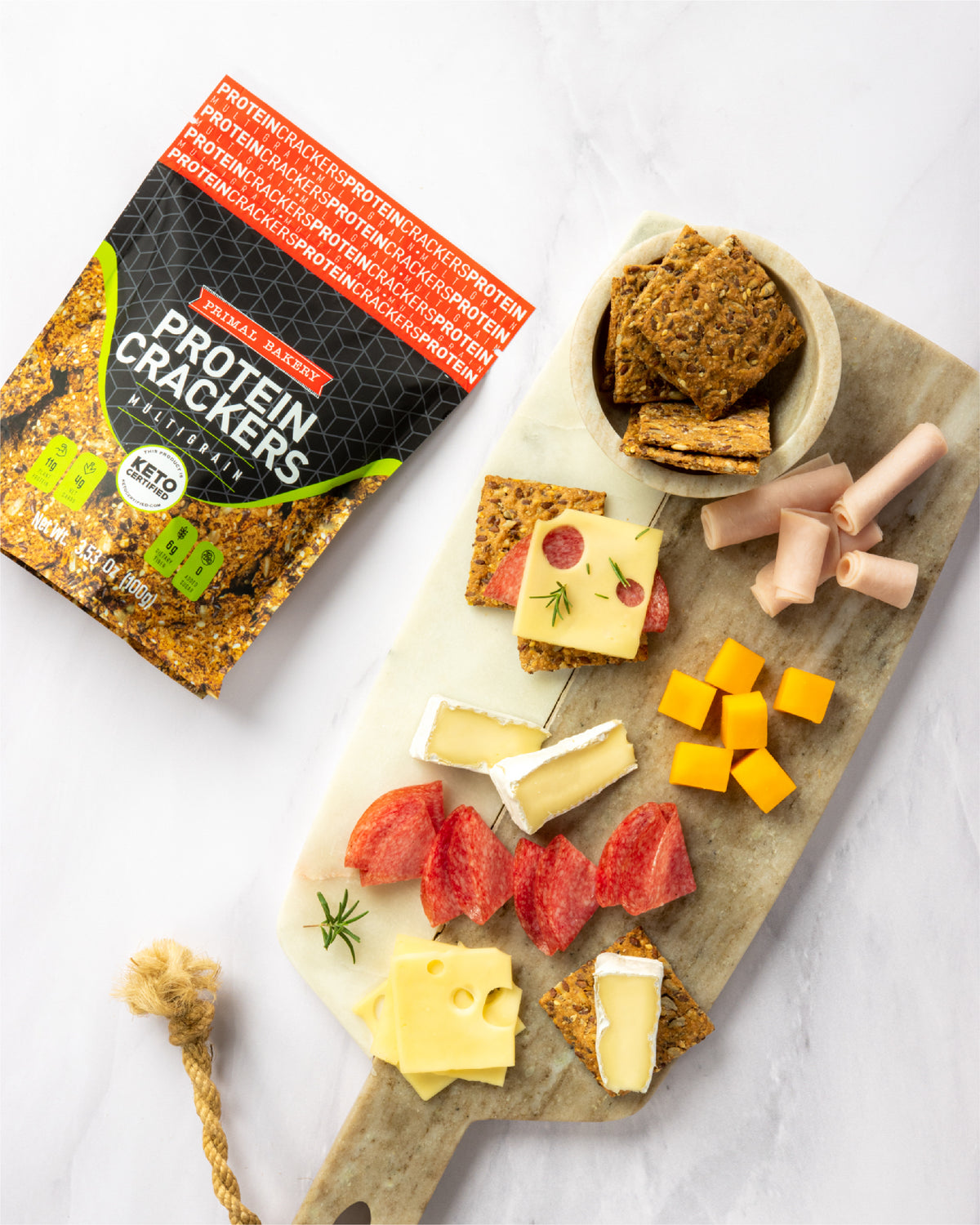 Primal Bakery Original Protein Crackers package beside a snack board with crackers, cheese, salami, and turkey.